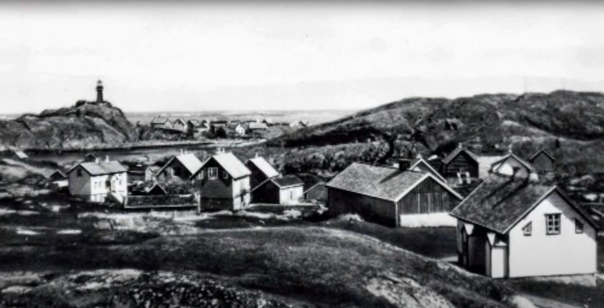 Historisk foto av Ona fiskevær med tettbygde hus plassert i le for vind og vær, og Ona Fyr på høyden i bakgrunnen.
